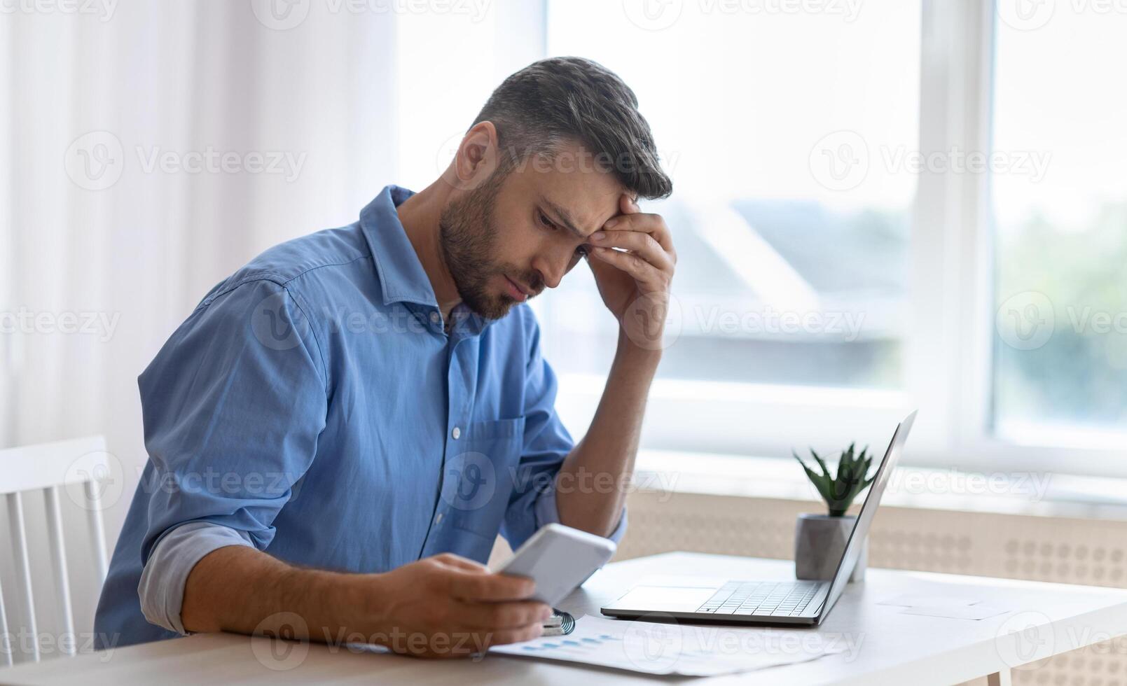 Worried male freelancer looking at smartphone screen while working at home office, received bad news, got scam message, thoughtful man touching forehead with concerned face expression, copy space photo
