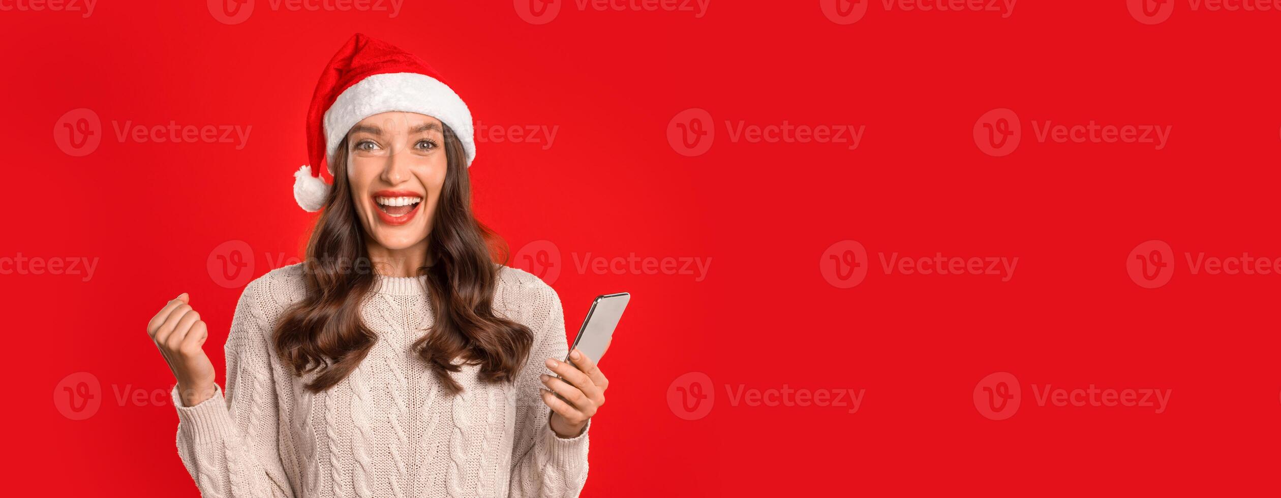 Young woman in Santa hat celebrates festive offer on phone, shaking fist raised in victory, against red studio backdrop. Portrait of joyful smartphone user gesturing yes holding gadget, copy space photo