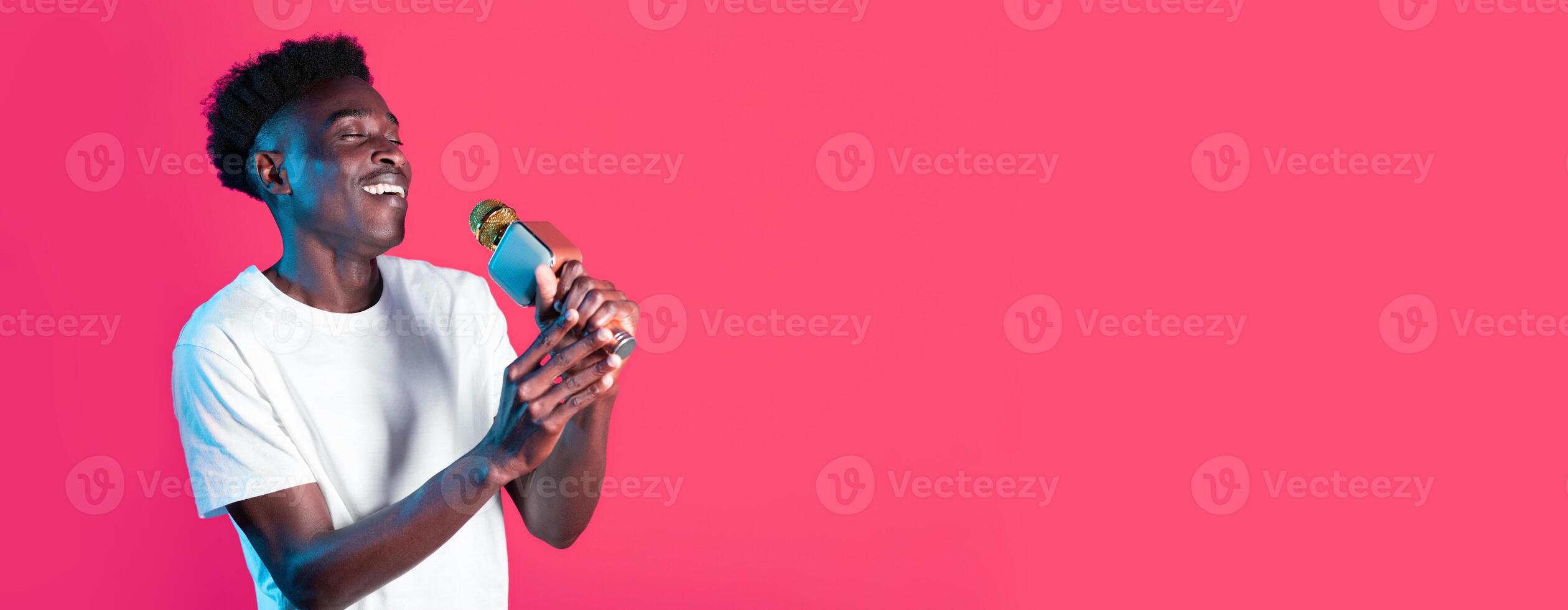 A young black man in a white t-shirt passionately sings into a microphone, smiling brightly. The vivid pink backdrop adds a lively touch to his energetic performance. photo