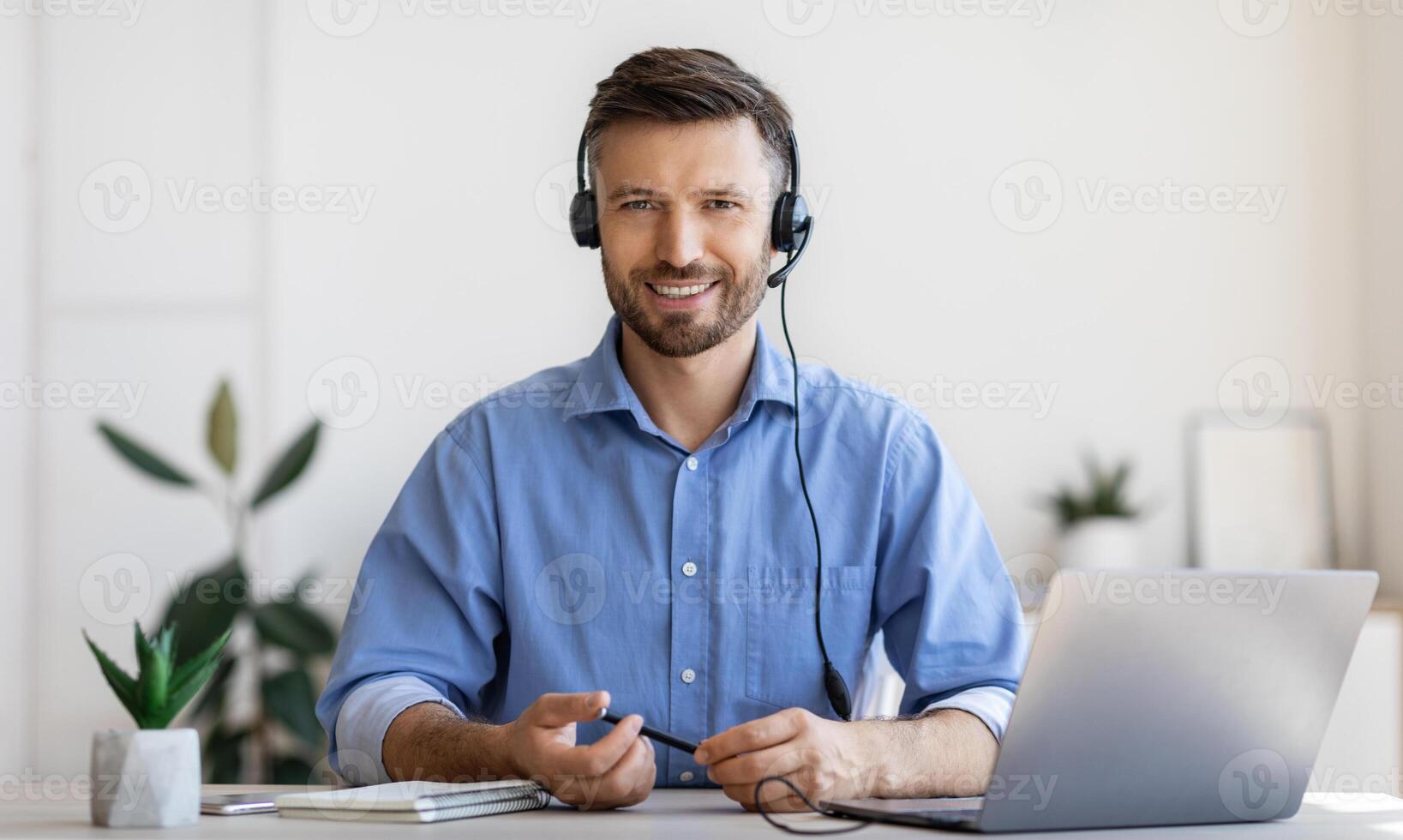 Portrait of smiling male customer service operator in headset posing at workplace, looking at camera, portrait of handsome hotline manager sitting at desk with laptop in white office, copy space photo