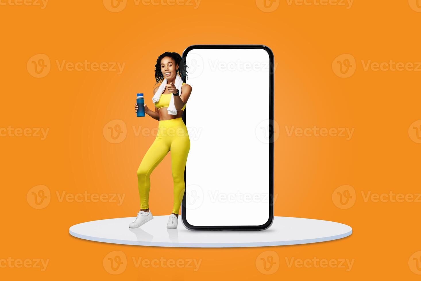 A fit woman stands confidently next to a large smartphone, holding a refreshing health drink. She wears bright yellow activewear, radiating energy and vitality in a vibrant setting. photo