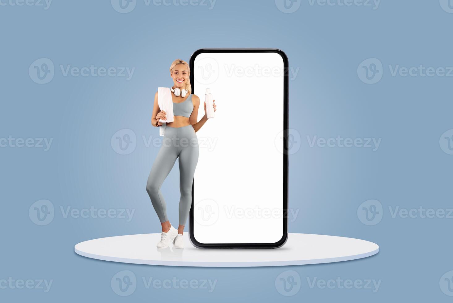 A fitness enthusiast stands confidently while holding a notebook and a drink. She is positioned next to a large blank smartphone screen, set against a light blue background. photo