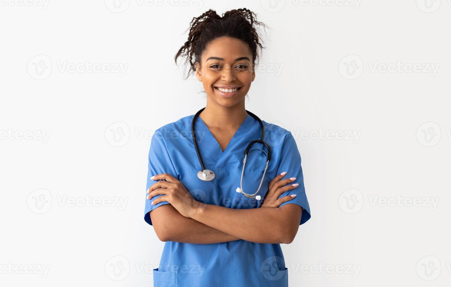 Portrait of positive female African American doctor or nurse posing with crossed arms and smiling looking at camera isolated over white studio background wall, lady wearing blue coat and stethoscope photo