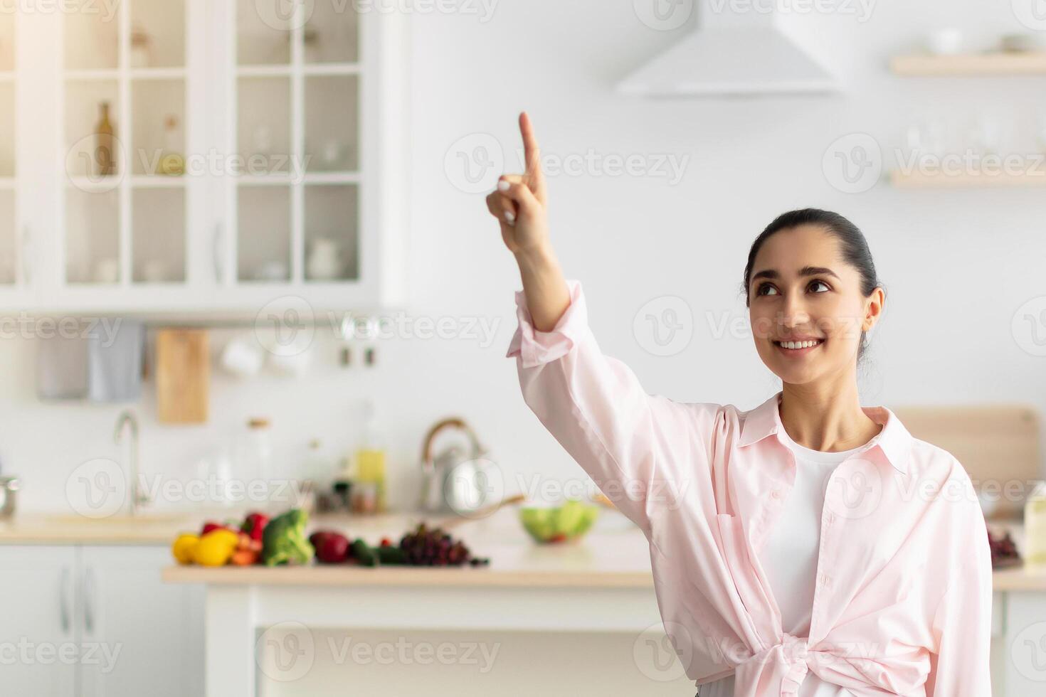 Modern Futuristic House Control System, IoT Technologies Concept. Portrait Of Smiling Millennial Lady Pointing Finger Up In The Air Pushing Button On Invisible Screen Standing In Kitchen Interior photo