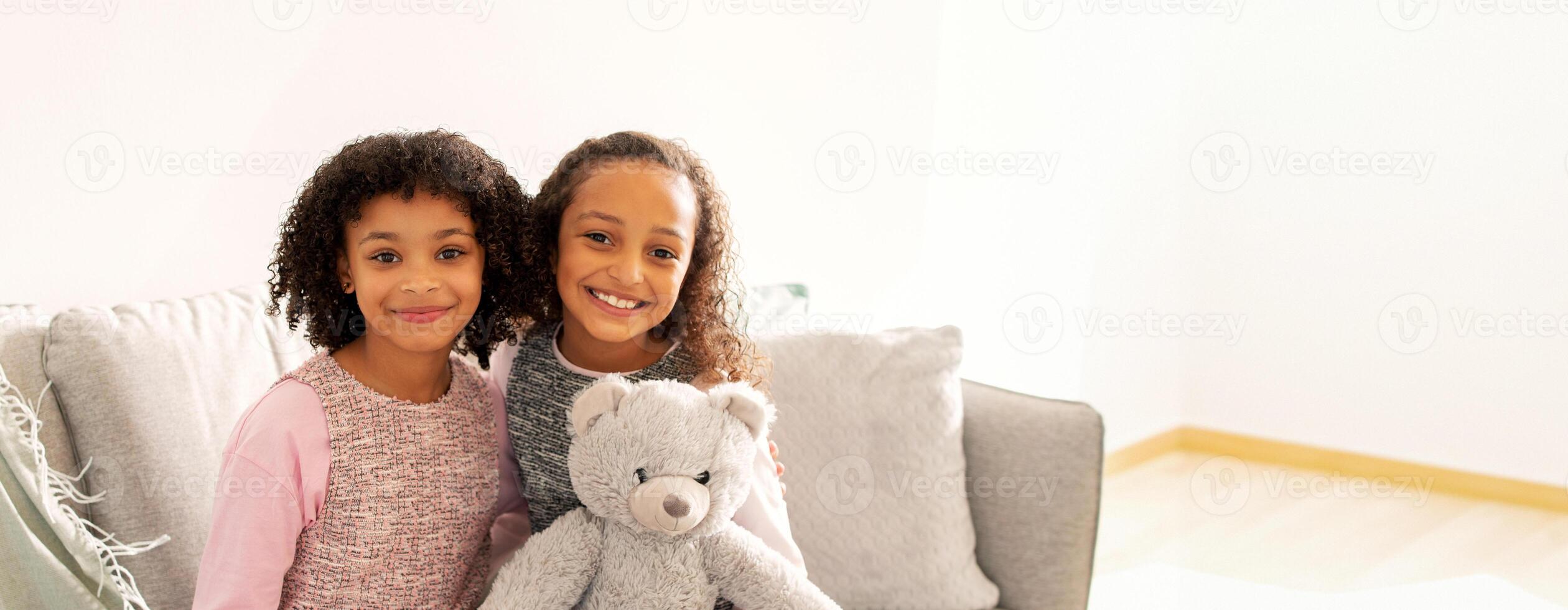 Two young black girls with curly hair are smiling widely while sitting on a sofa in a well-lit living room. One girl is holding a soft teddy bear, creating a joyful atmosphere. photo