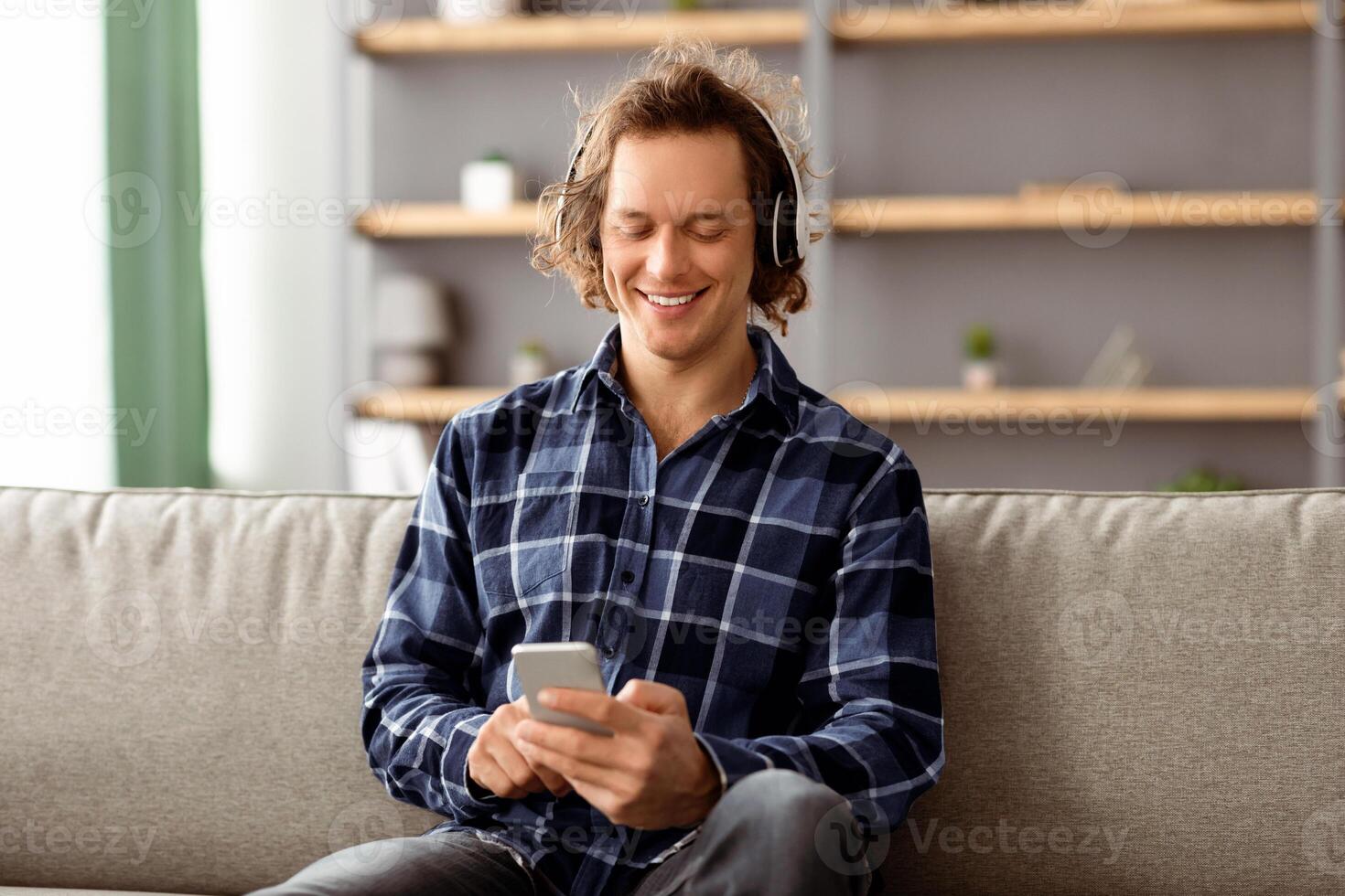 Happy Man Listening To Music On Phone Online Enjoying Good Sound Wearing Wireless Headphones Sitting On Couch At Home. Guy Relaxing Using Musical Application Browsing Favorite Song On Smartphone photo