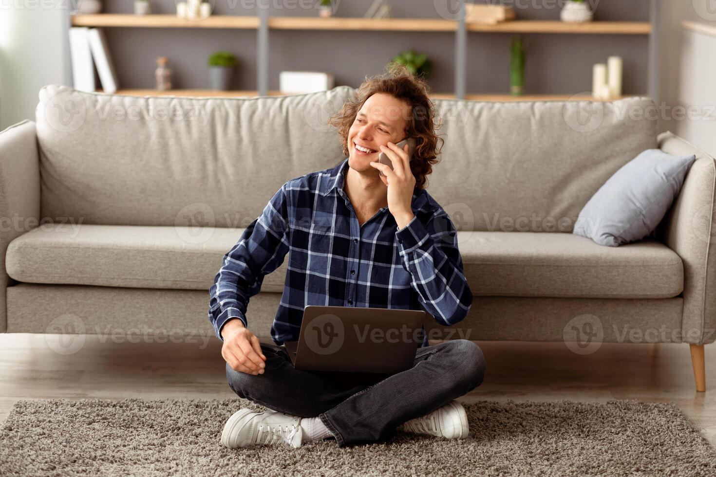 Mobile Communication. Cheerful Guy Talking On Smartphone Sitting With Laptop On Floor At Home, Working Remotely Online. Man Calling Speaking By Phone Having Pleasant Conversation photo