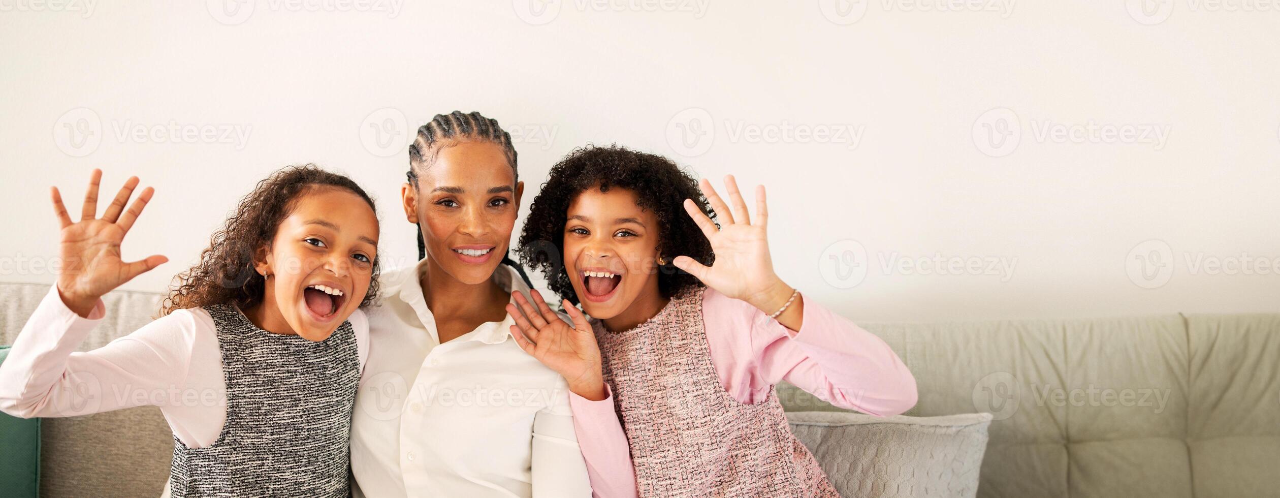 A black mother sits on the couch with her two daughters, all smiling and playfully waving their hands. They're enjoying a moment of happiness together in a cozy living room setting. photo