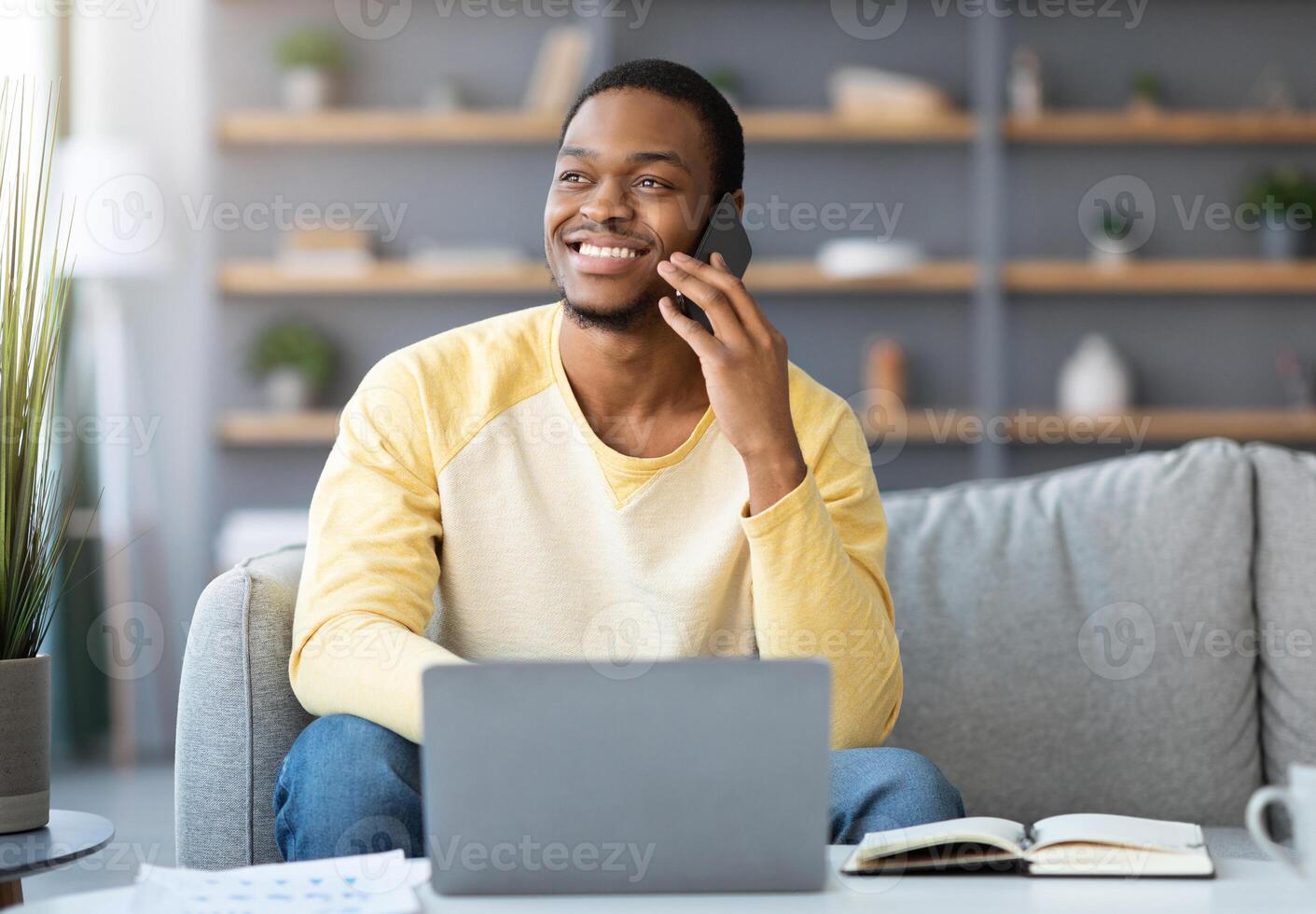 Joyful black guy freelancer having phone conversation while working from home, using laptop, smiling and looking at copy space. Young african american businessman talking with clients on mobile phone photo