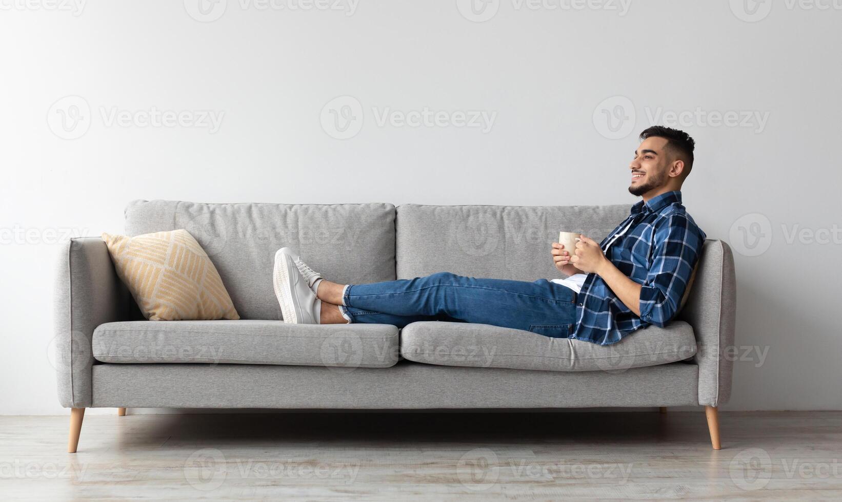 Rest Concept. Happy Arab guy drinking coffee sitting on comfortable couch at home in living room. Cheerful casual man relaxing on sofa, enjoying weekend free time or break from work, full body length photo