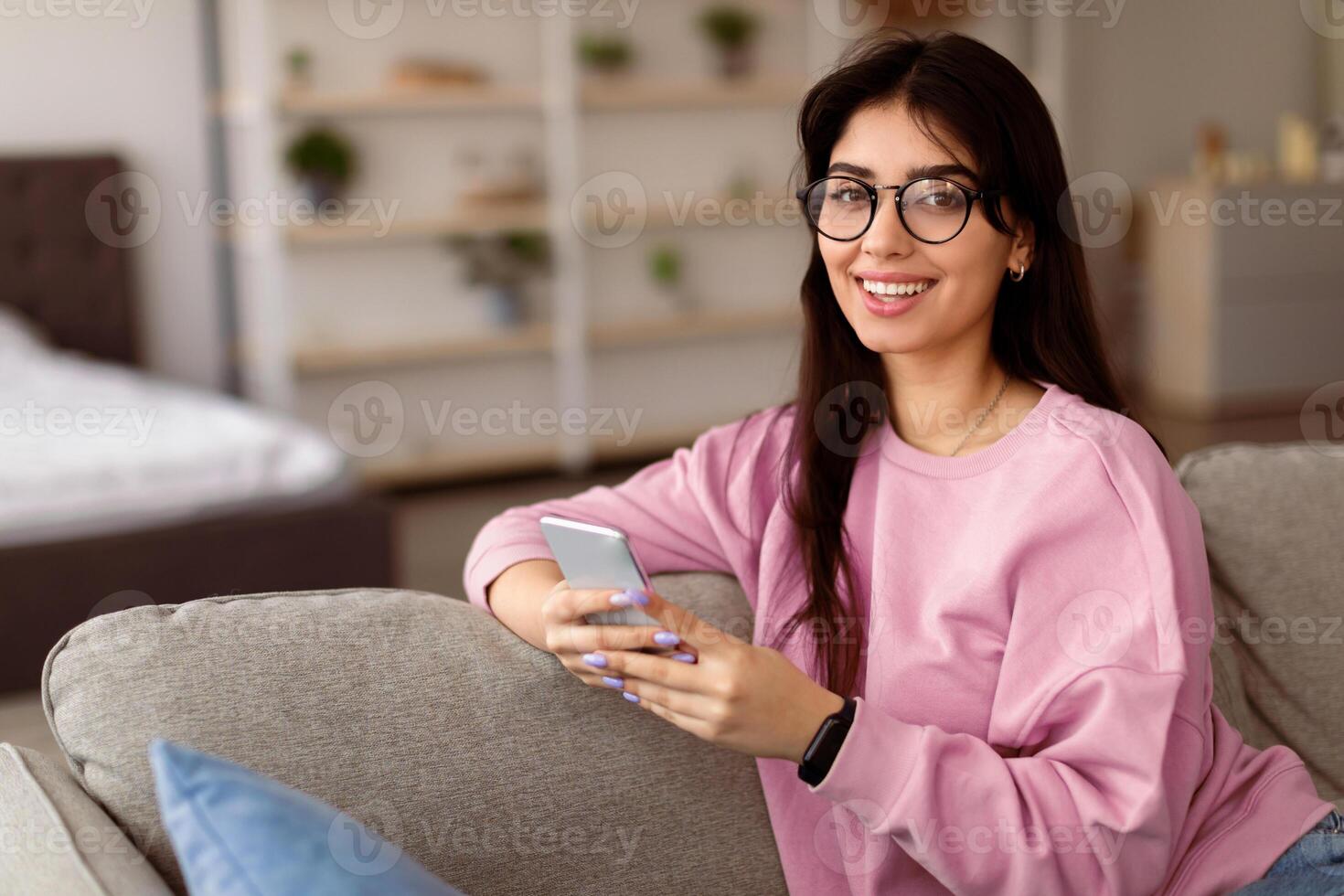 People And Technology Concept. Portrait of excited smiling young woman in eyeglasses using smartphone sitting and leaning on the couch, looking at camera. Lady chatting online, selective focus photo
