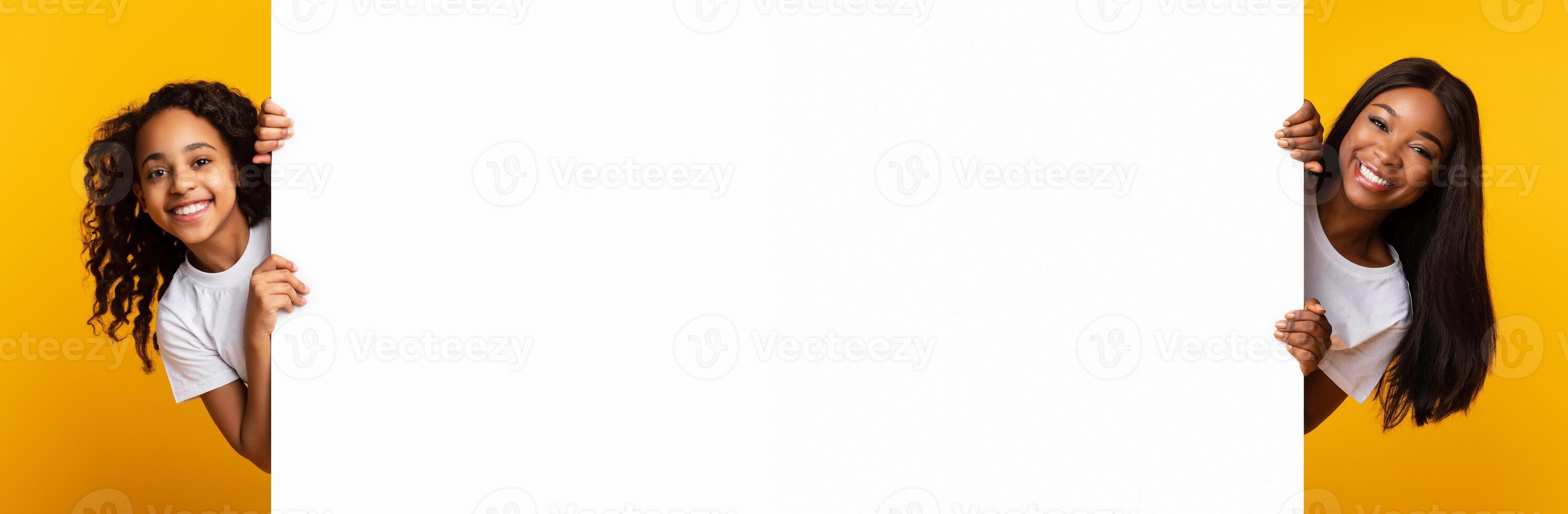 Promotion Billboard Concept. Cheerful African Woman Mom And Girl Holding White Advertisement Board, Standing Together On Yellow Background, Looking At Camera Peeping Out The Two Sides Of Sign Board photo