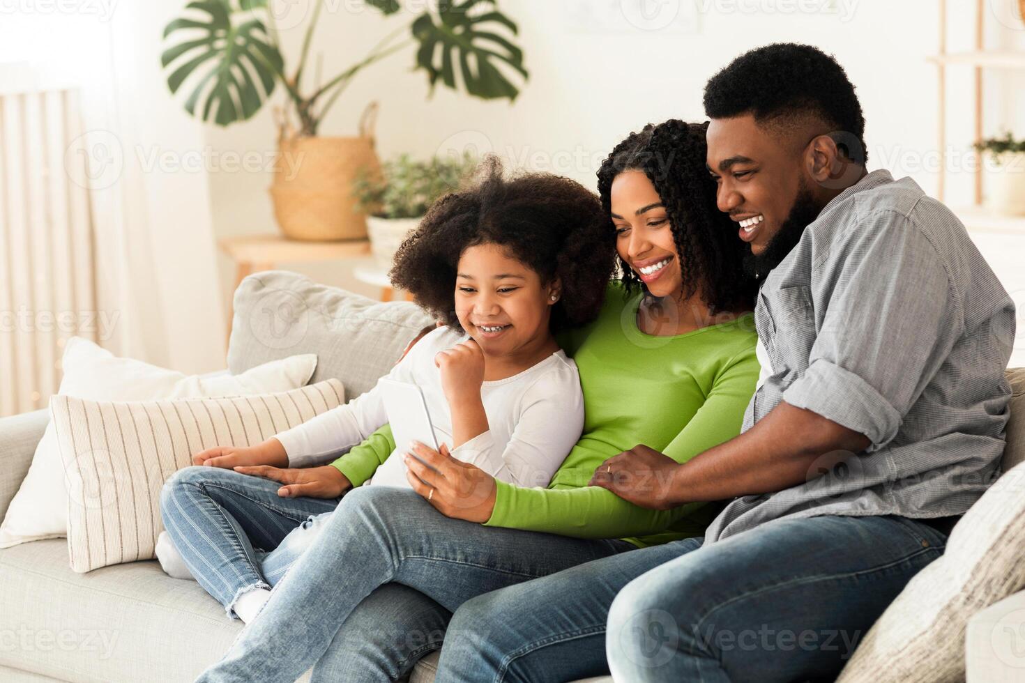 Happy black father, mother and their little daughter relaxing with smartphone at home together, sitting on couch, watching videos or making call photo