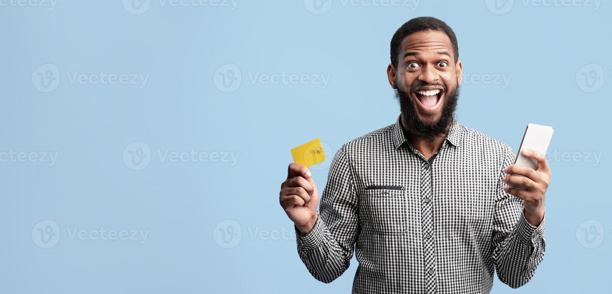 Mobile Payment Concept. Excited African American Man Holding Credit Card And Smartphone Posing Standing Isolated Over Blue Studio Background. Joyful Male Model Making Online Purchase photo