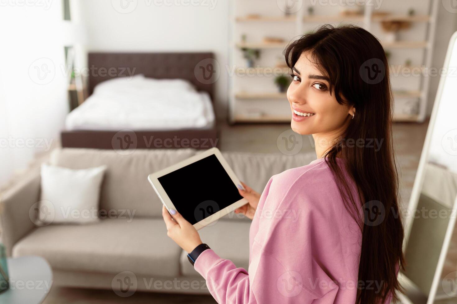Portrait of smiling young woman holding digital tablet with black empty screen for mockup template, looking back at camera, standing in modern living room. Technology, Automation, Smart Home Concept photo