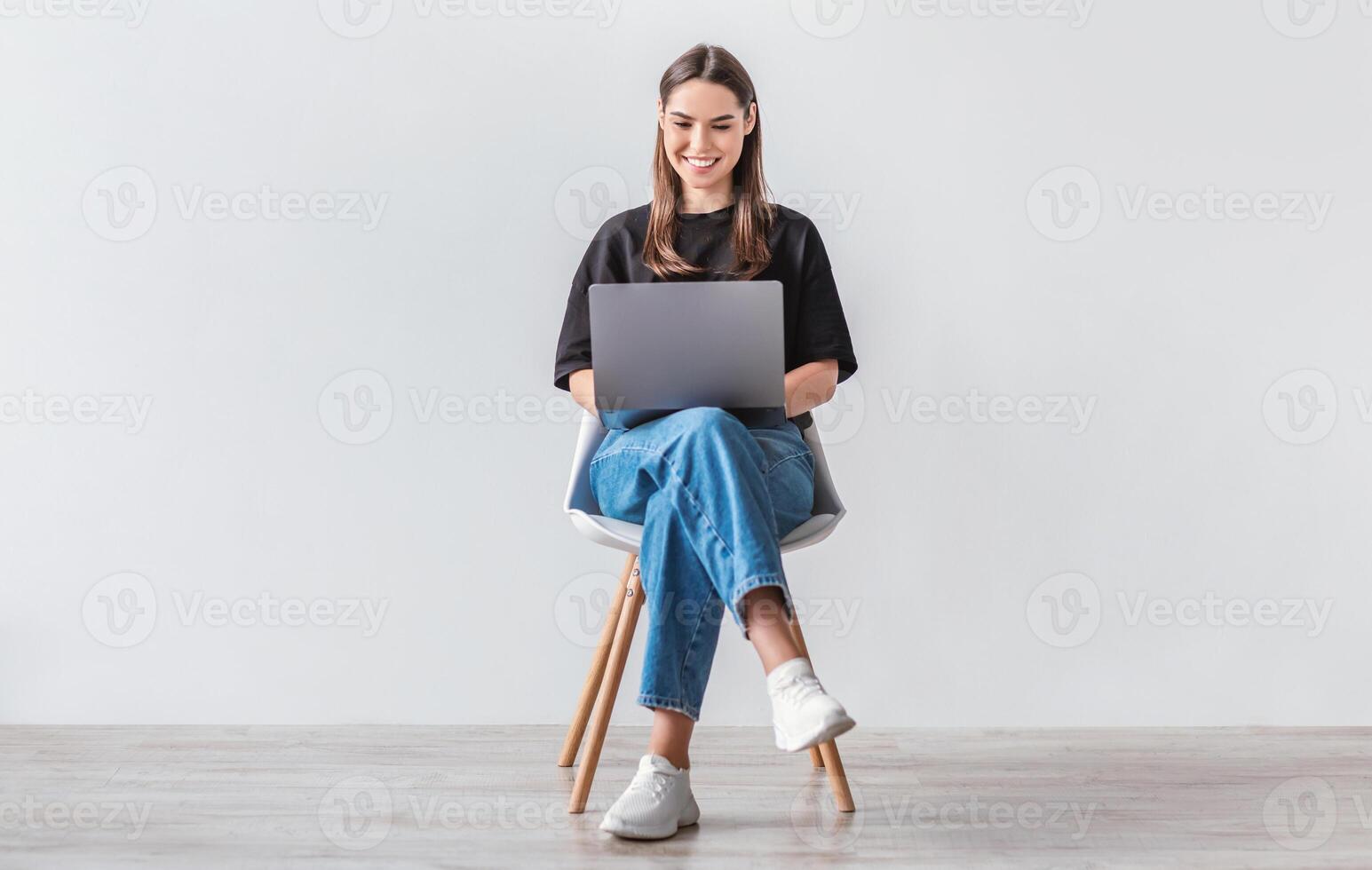 Happy young woman working online, sitting on chair and using laptop against white studio wall, full length. Positive Caucasian lady surfing internet, watching webinar on portable computer photo