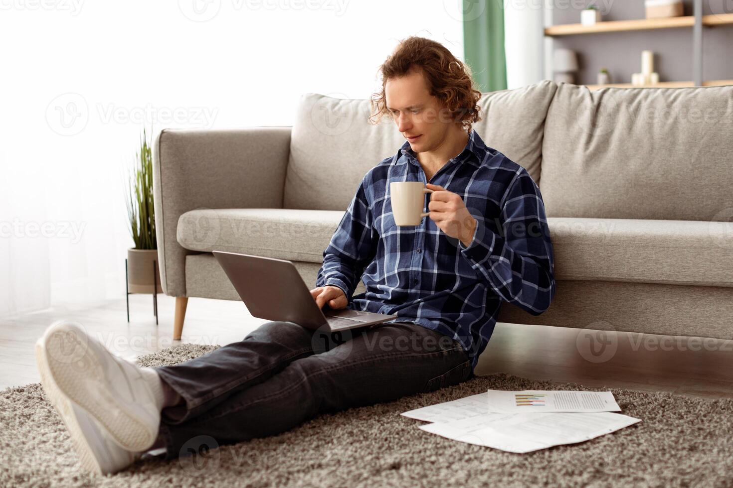 Freelance Work. Serious Guy Using Laptop Computer Working Online And Drinking Coffee Sitting On Floor At Home. Male Freelancer Browsing Internet Indoors. Internet And Technology, Remote Job photo