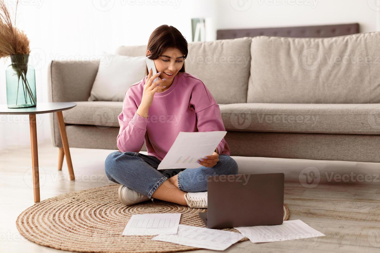 Business, Technology And Communication Concept. Young smiling woman making phone call while looking at document with graphs, busy employee sitting on floor at home office with laptop computer photo