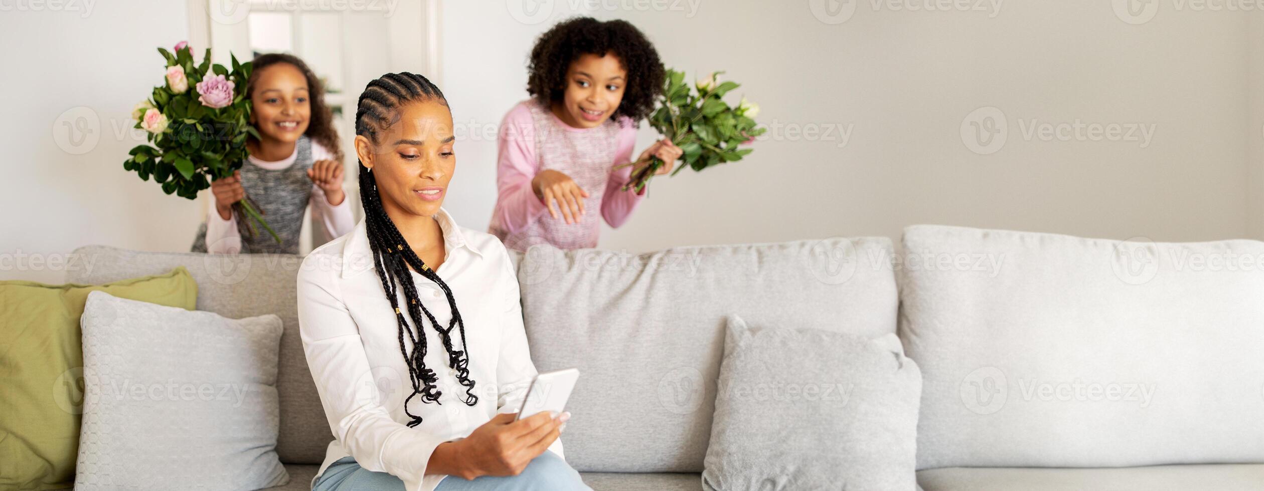 A black mother sits on a couch, focused on her phone, while her two daughters sneak up behind her, holding flowers. The cozy living room setting adds warmth to the joyful moment of surprise. photo