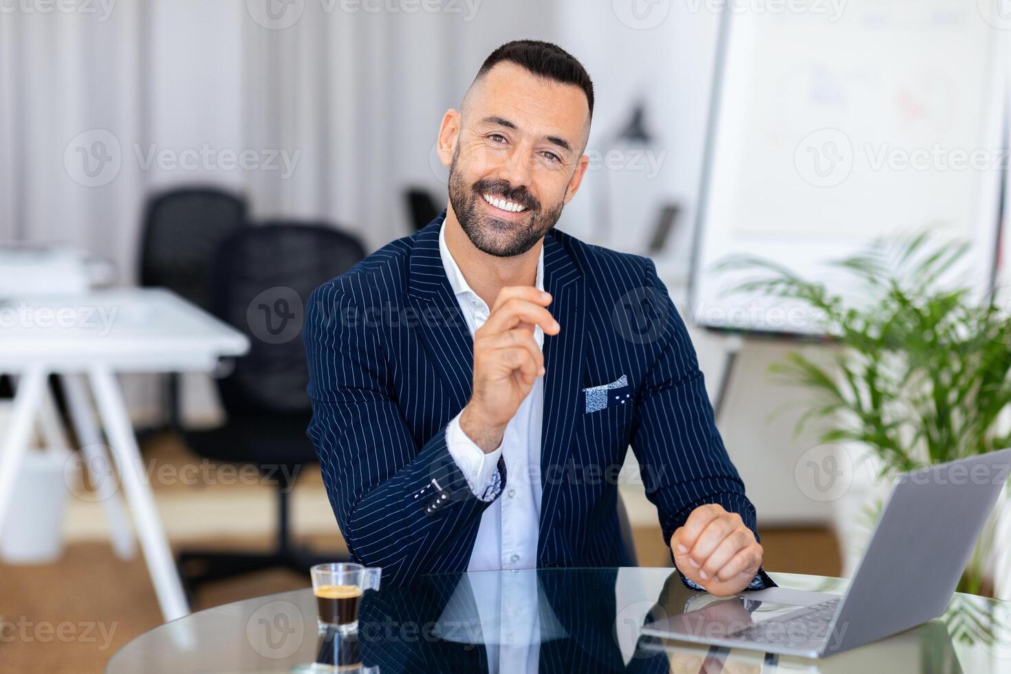 A confident man in a pinstripe suit sits at a sleek office desk, smiling and holding a pen. The environment is contemporary, with plants and minimalistic furniture in the background. photo
