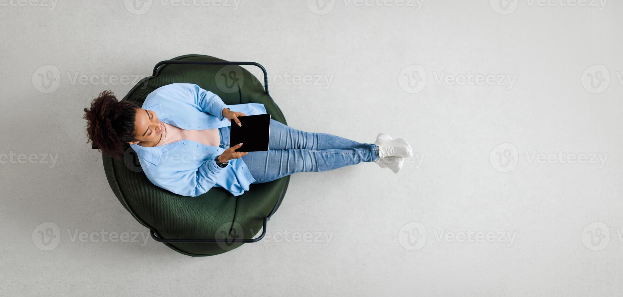 People And Modern Technology. Above top view of young smiling black lady holding tablet sitting on bean bag in living room, watching . Cheerful lady surfing internet, free copy space, panorama photo