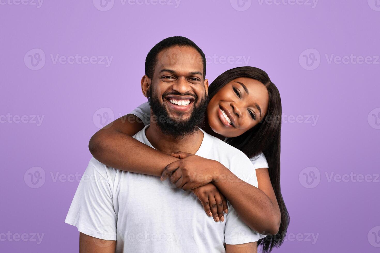 Tenderness Concept. Portrait of charming cute black lady hugging her man from behind, enjoying time together, standing isolated over yblue studio background, posing and looking at camera photo
