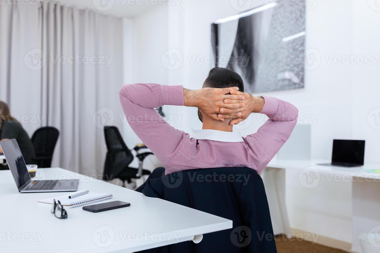 A man in a casual shirt leans back in his chair, hands clasped behind his head, enjoying a moment of relaxation in a contemporary office setting, surrounded by desks and laptops. photo