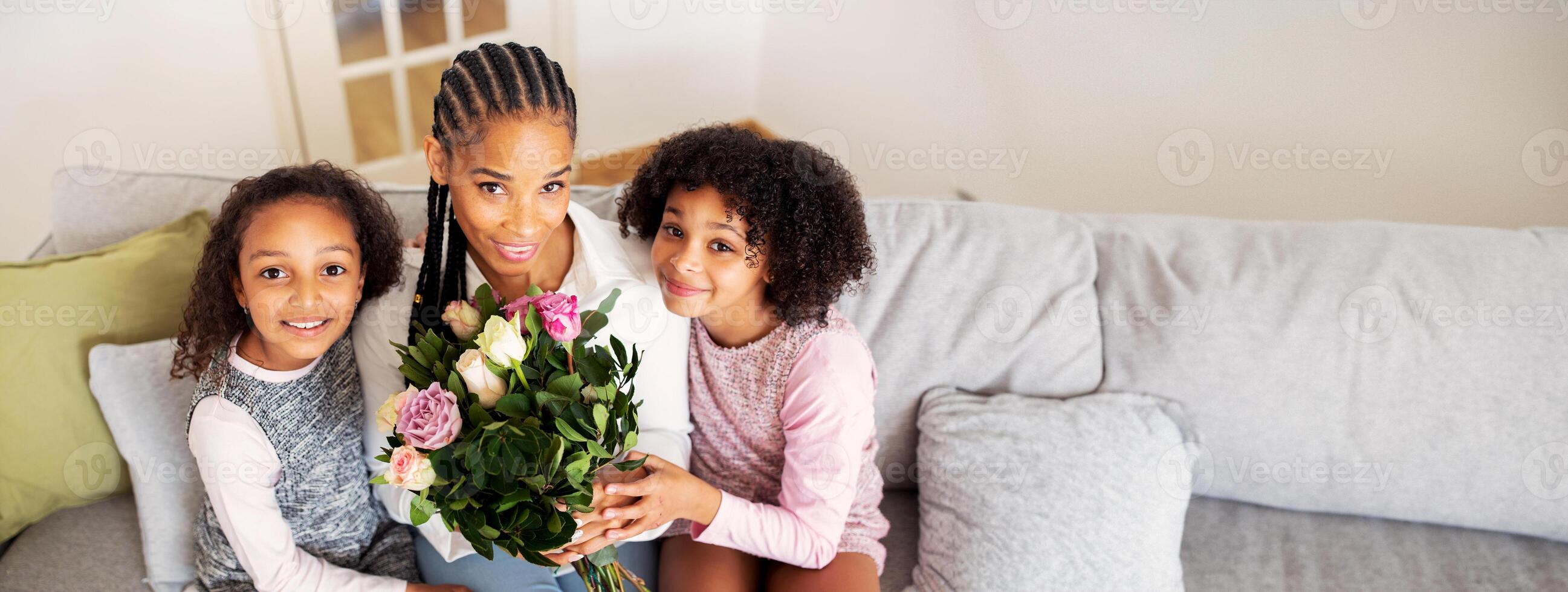A black mother sits on a couch surrounded by her two daughters, all smiling warmly. She holds a bouquet of flowers, symbolizing a joyful occasion, enhancing their bonding time. photo