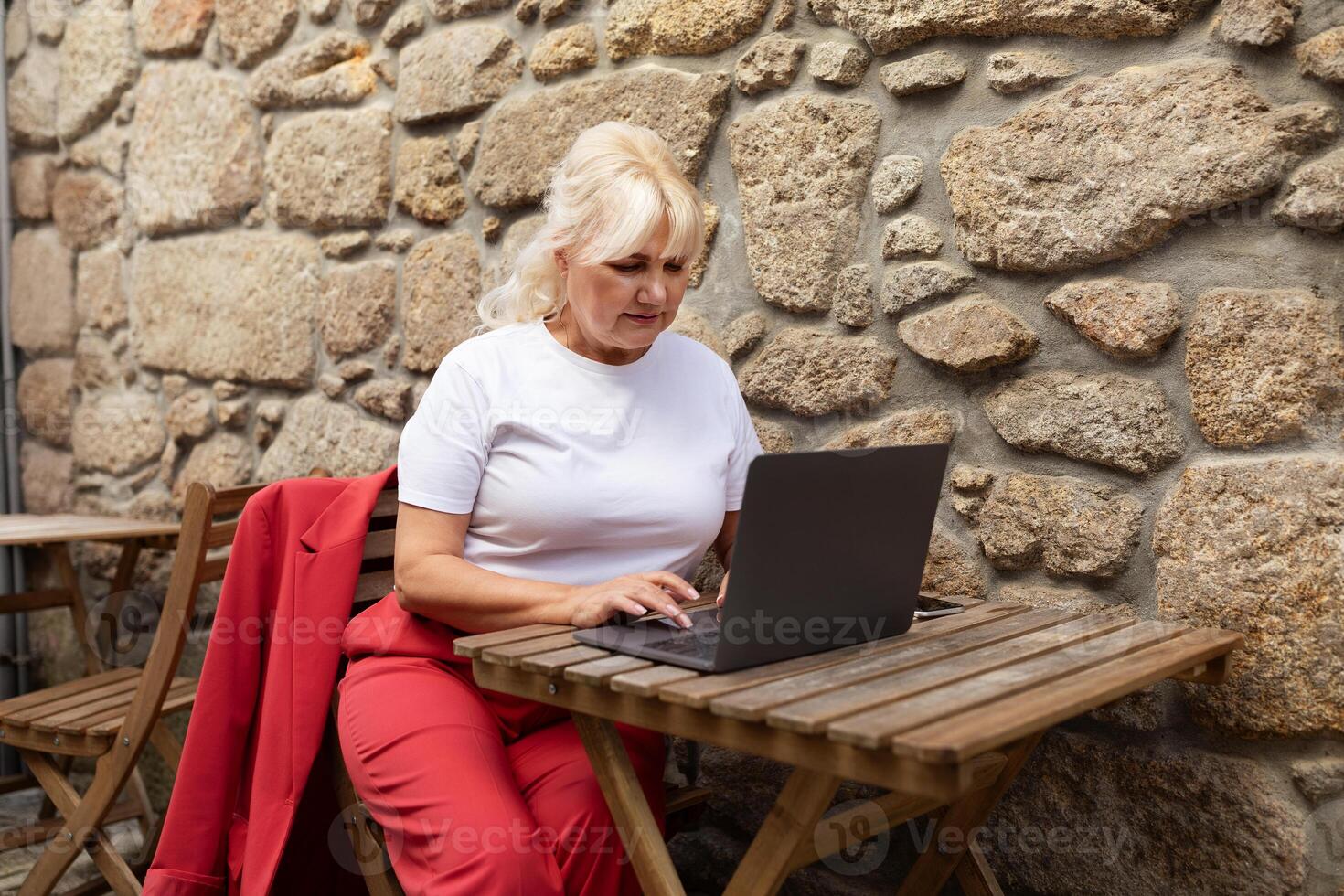 A woman is focused on her laptop while sitting at a wooden table in a cozy cafe. The rustic stone wall adds charm to her work environment, creating a serene outdoor atmosphere. photo