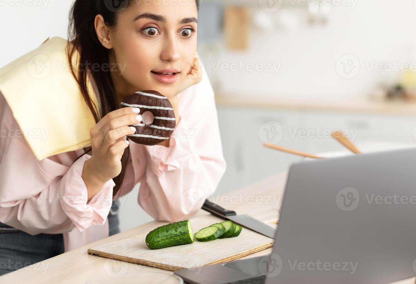 Exciting Movie. Amazed surprised young lady watching film or reality show standing in kitchen indoors at home, using laptop computer while cooking, eating sweet doughnut, leaning on table photo