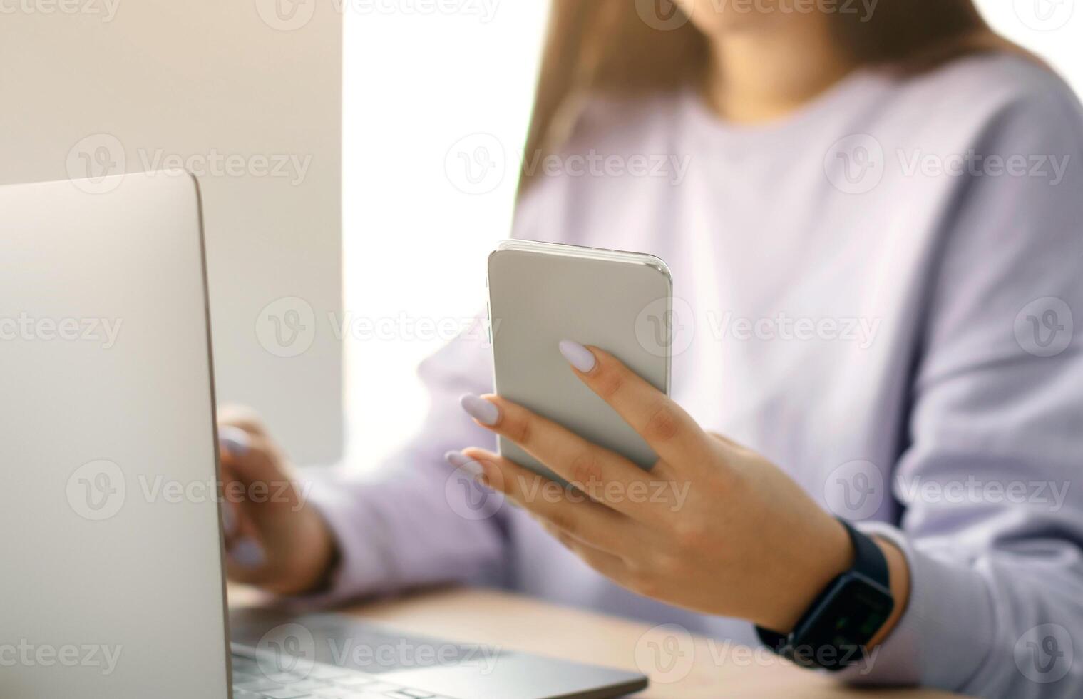 People And Technology. Closeup cropped view of smiling unrecognizable lady holding her smartphone and using modern laptop computer, reading business text message, sitting at desk, selective focus photo