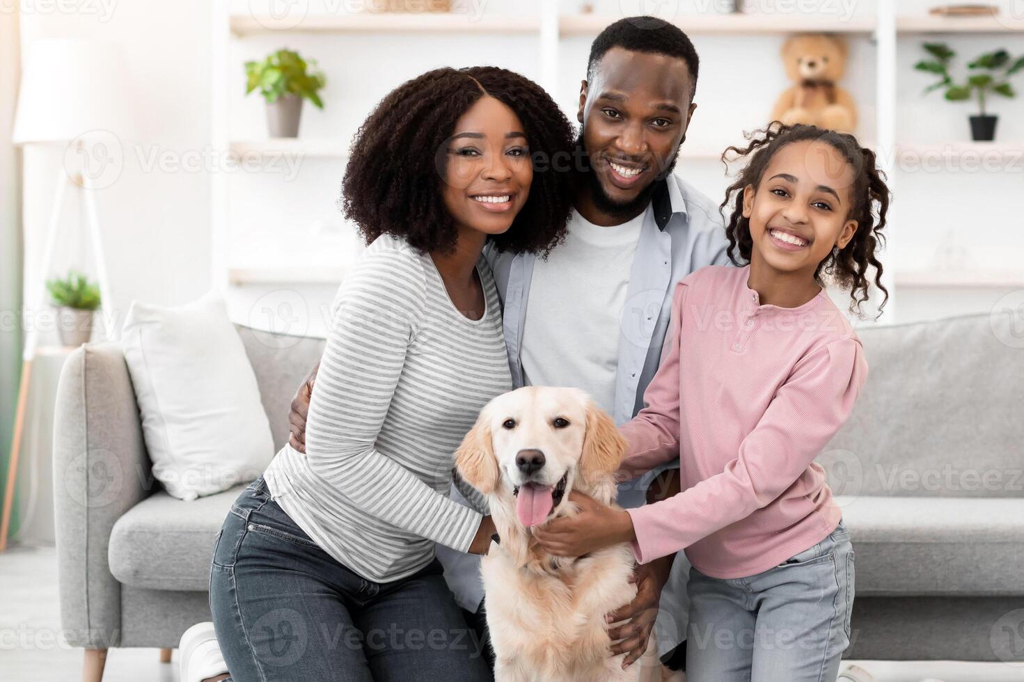 Happy Loving Family. Portrait of smiling African American man, woman and girl hugging and stroking their happy dog sitting on the floor carpet in living room at home, posing and looking at camera photo