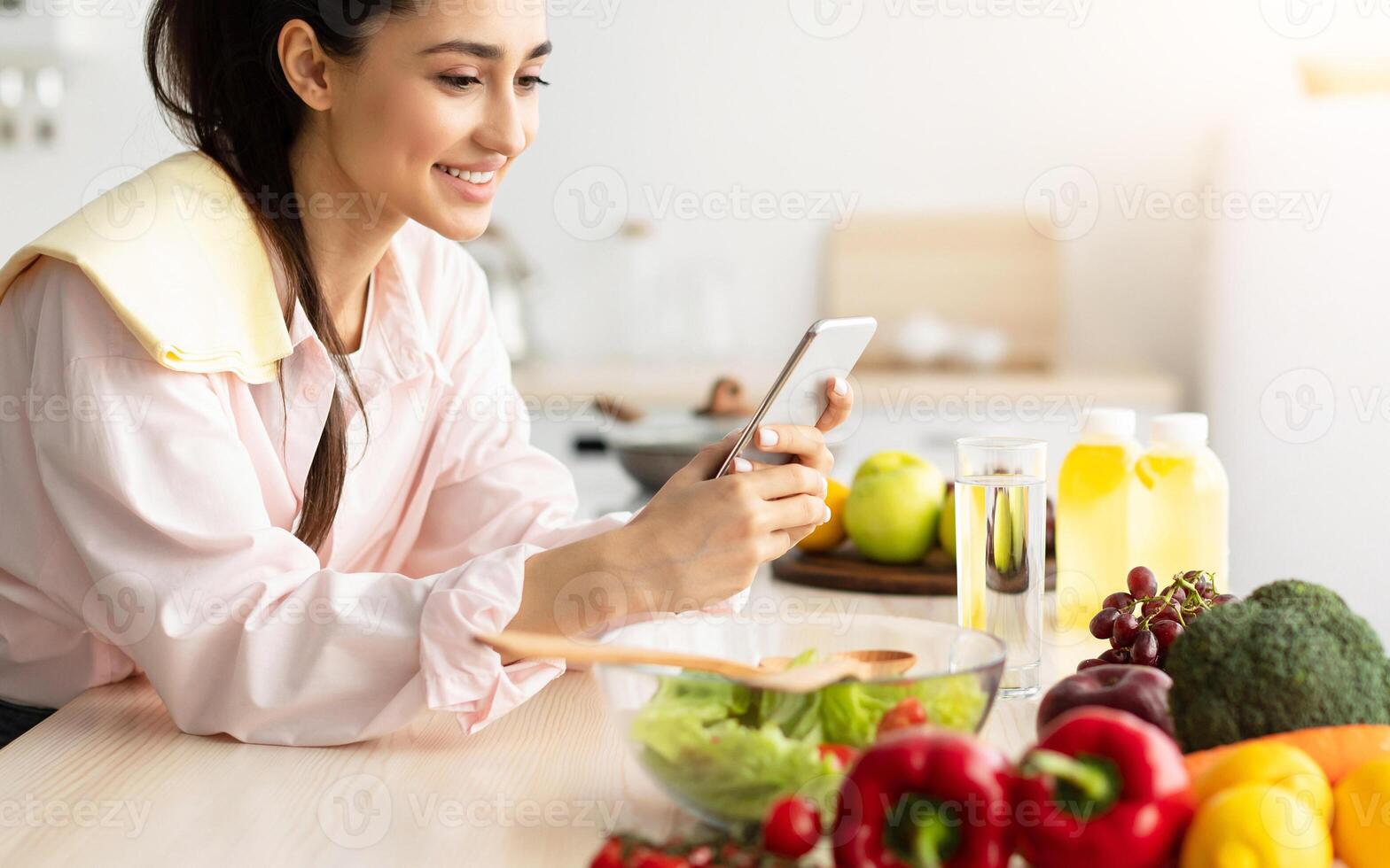 Smiling young woman using cellphone while cooking dinner in kitchen at home, cheerful casual female leaning on table, reading text messages on her cell phone and smiling, free copy space photo