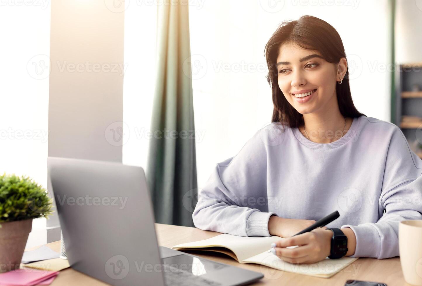 Home Office And Telecommuting Concept. Potrait of smiling casual woman sitting at desk, using laptop. Female writing in notebook, taking notes, working or studying remotely, free space photo