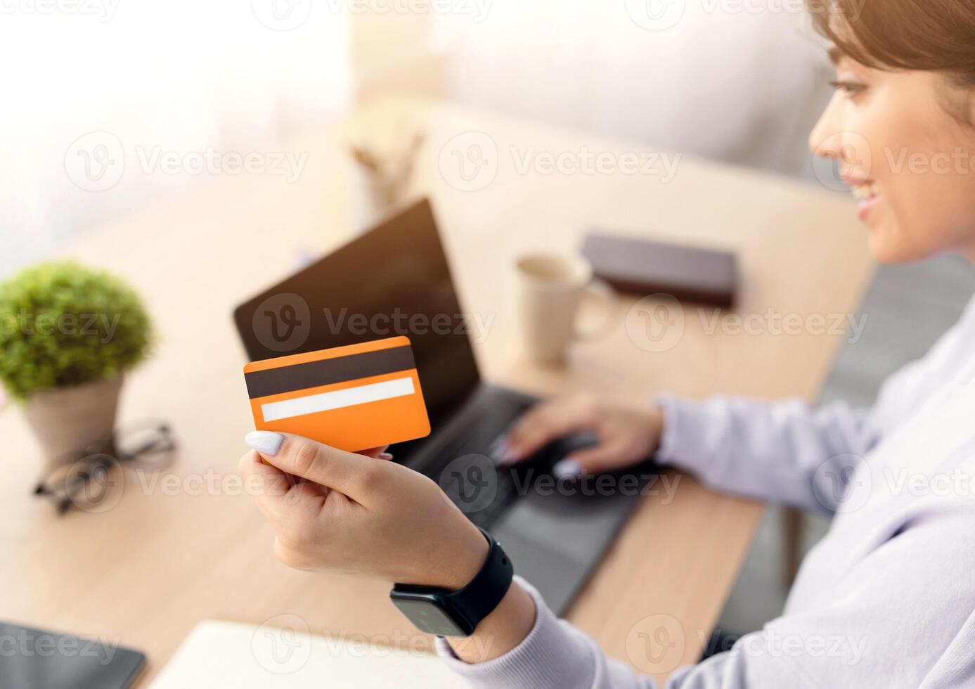 Online Shopping Concept. Closeup of smiling young lady holding and showing golden credit card, sitting at desk with laptop computer at home, making financial transaction, selective focus photo