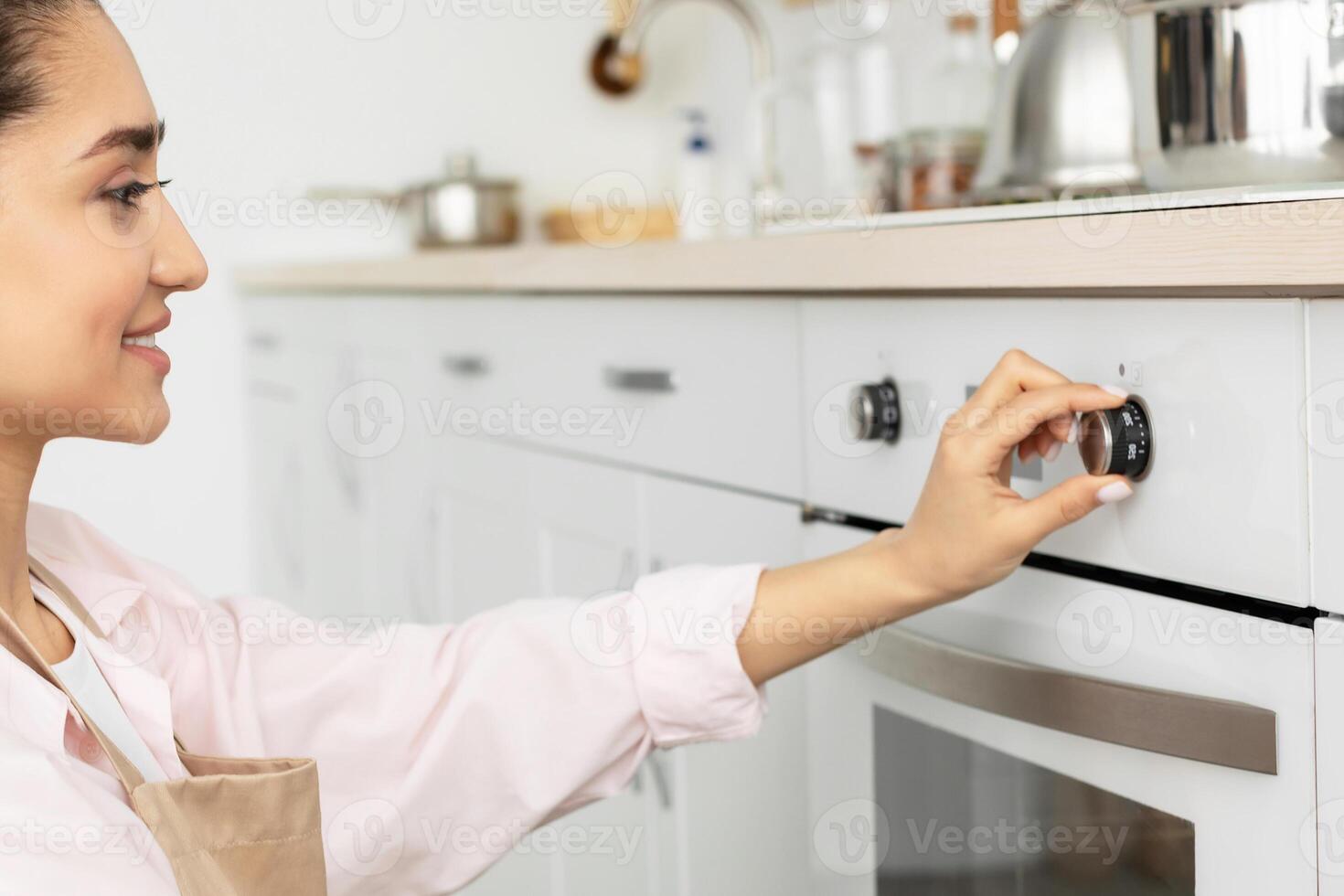 Household Concept. Closeup side view portrait of millennial cheerful woman in apron using stove in modern kitchen at her new apartment, holding and turning knob, controlling temperature of the burner photo