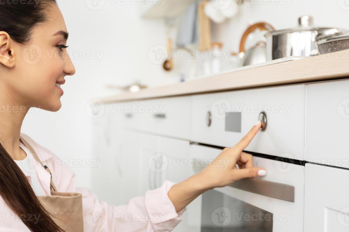 Closeup side view profile portrait of young smiling woman in apron using stove in modern kitchen at home, touching and pressing switch button, controlling temperature of the burner, preparing meal photo