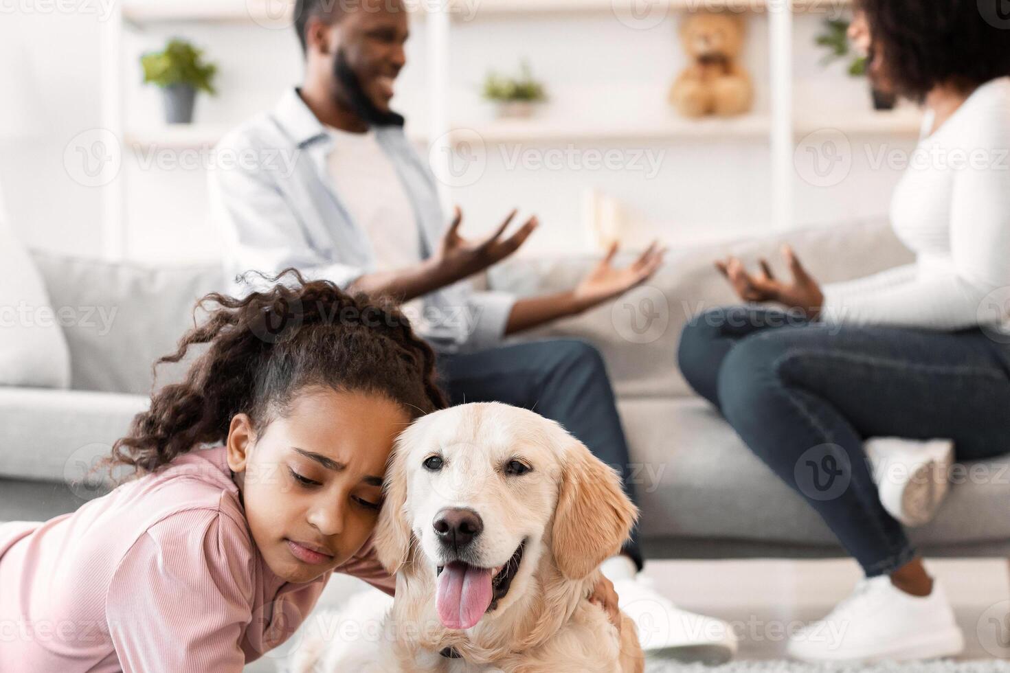 Divorce, Breakup And Children. Portrait of upset African American daughter hugging doog and lying on floor while her angry parents fighting in the background, depressed child feeling lonely photo