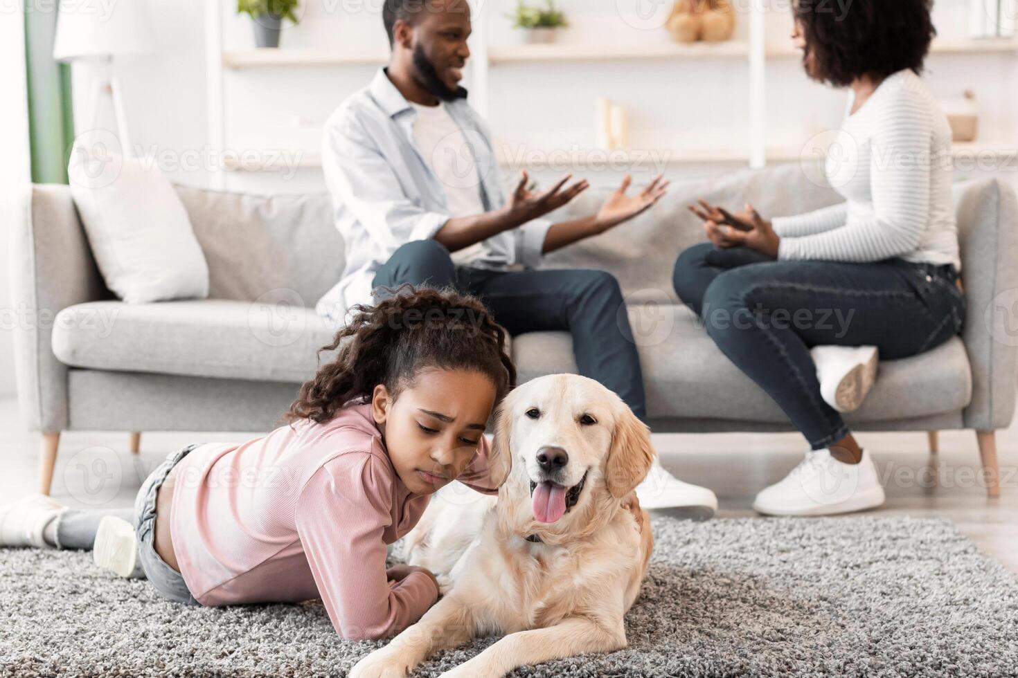 Family Conflicts. Sad little black girl hugging dog while her parents arguing in the background, upset child hearing quarrel and lying on floor carpet, selective focus on stressed kid and pet photo