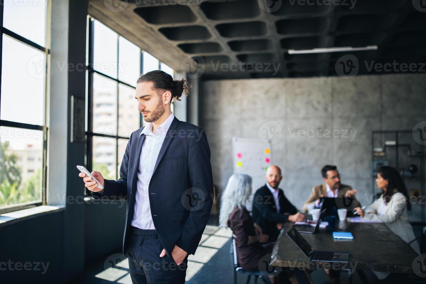 A man in a suit stands by a window, attentively checking his phone while colleagues engage in a discussion at a conference table. The setting is a contemporary office filled with natural light. photo