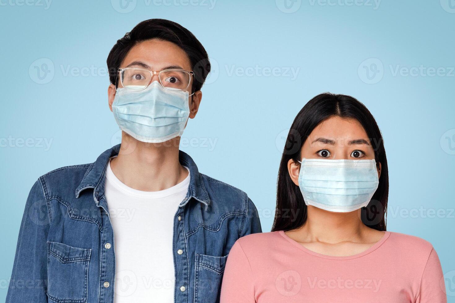 Coronavirus Concept. Frightened asian couple posing in protective medical face masks, standing over light background in studio, empty space photo