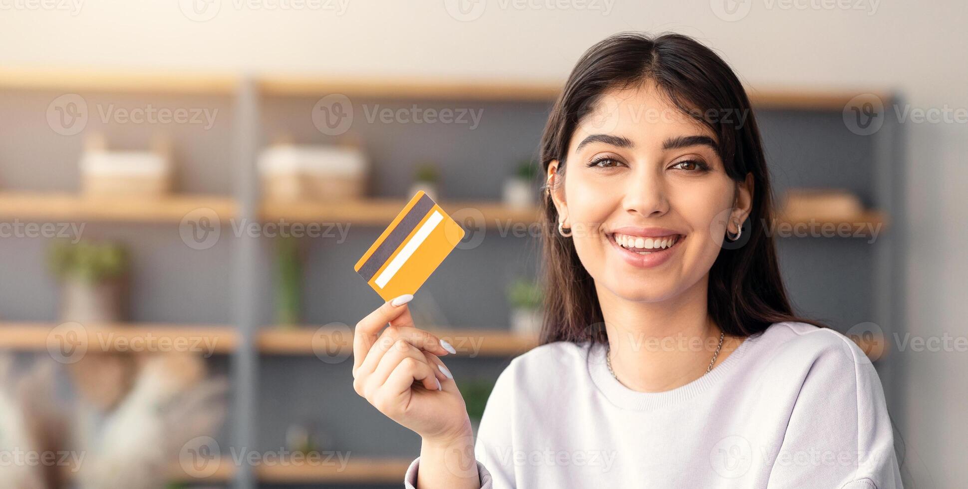 Making Financial Transaction. Portrait of smiling young lady holding and showing golden credit card, sitting at desk with laptop computer at home, looking at camera. Online Money Transfer Cocnept photo
