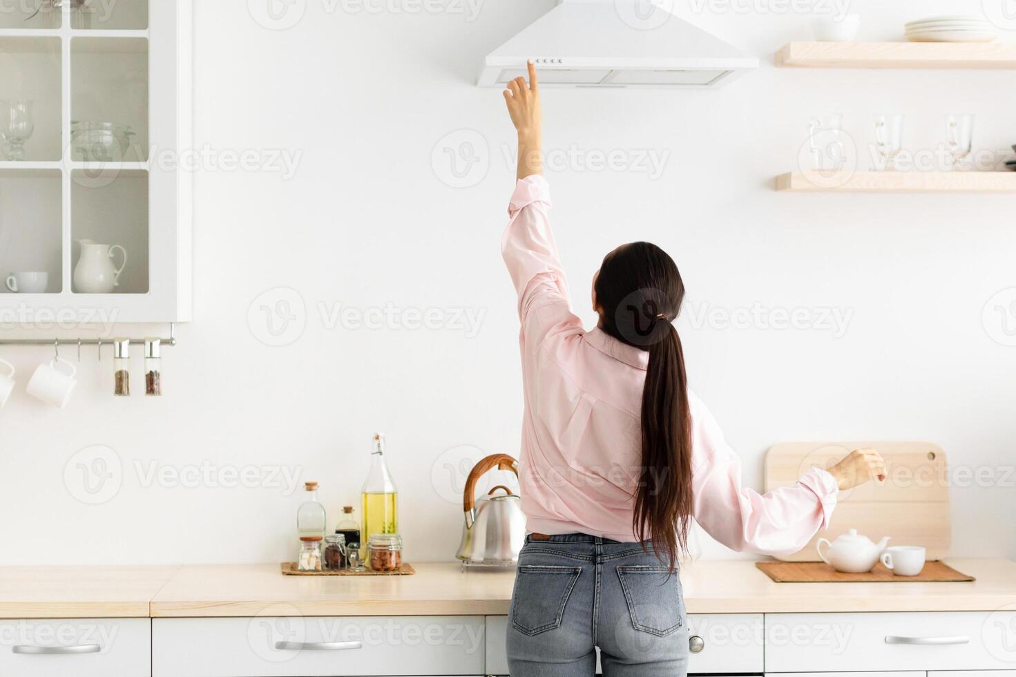 Rear back view of young millennial housewife choosing and selecting mode on range exhaust hood, pushing button on mechanical fan above the stove, standing in modern kitchen at home photo