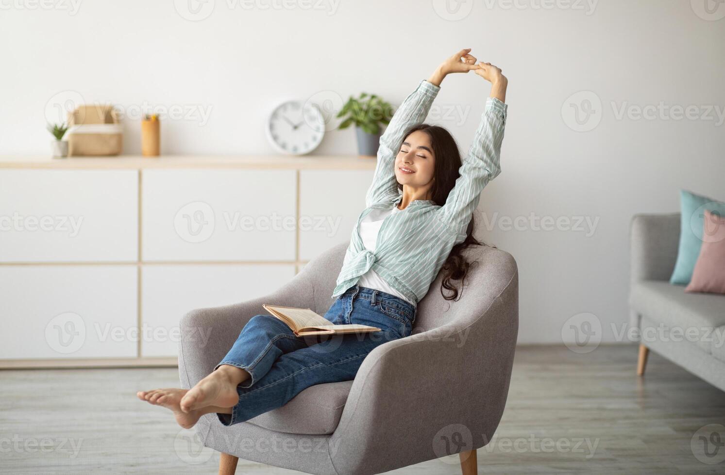 Gorgeous Indian woman stretching in comfy armchair while reading indoors. Attractive young lady with open book, enjoying lazy morning, having peaceful weekend. Stay home hobbies photo