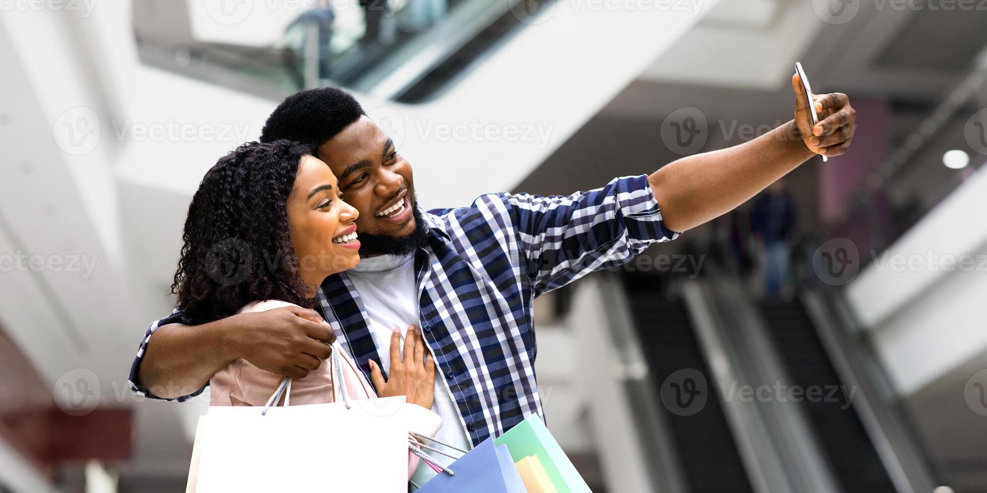 Positive Black Spouses Shopping In Department Store And Taking Selfie On Smartphone, African Couple Having Fun In Mall, Enjoying Doing Purchases Together, Carrying Colorful Shopper Bags, Free Space photo