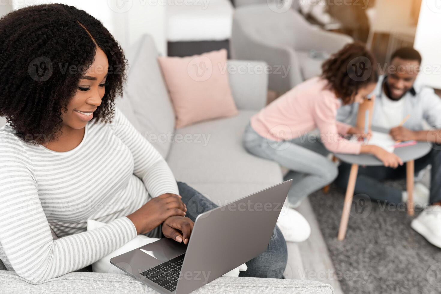 Smiling Black Woman Working Online On Laptop, Sitting On Couch With Computer Reading Email, Enjoying Freelance Work. Young Father Sitting On Floor Spending Time With Daughter Drawing On Table photo