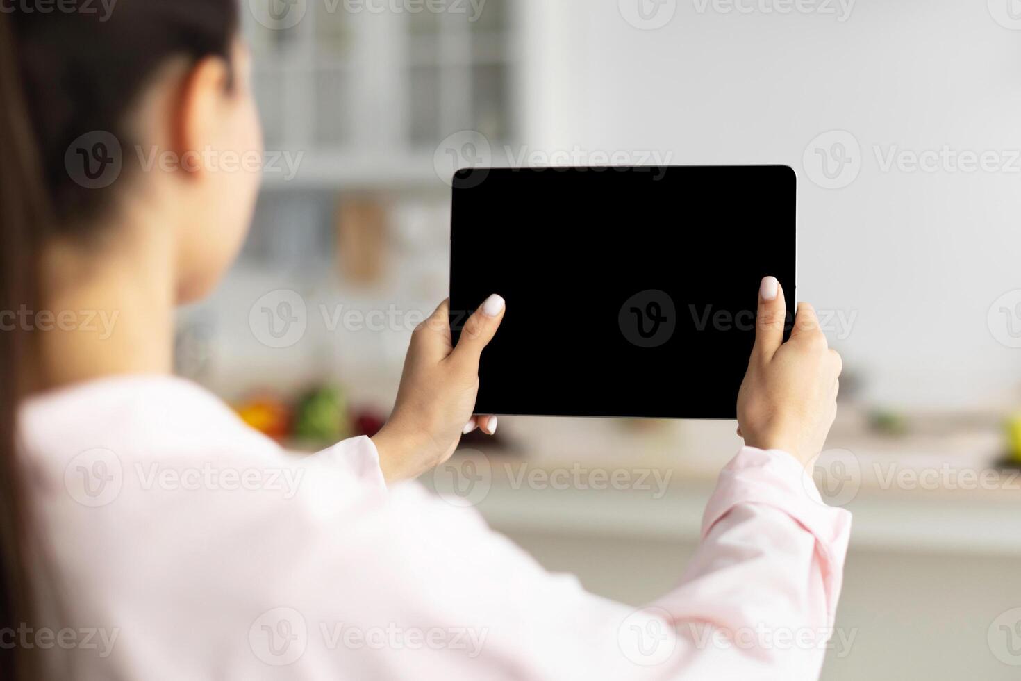 Over The Shoulder View Of Unrecognizable Woman Using Digital Tablet With Empty Screen Standing In Kitchen At Home. Application Or Internet Website Advertising, Mock Up, Free Space, Selective Focus photo