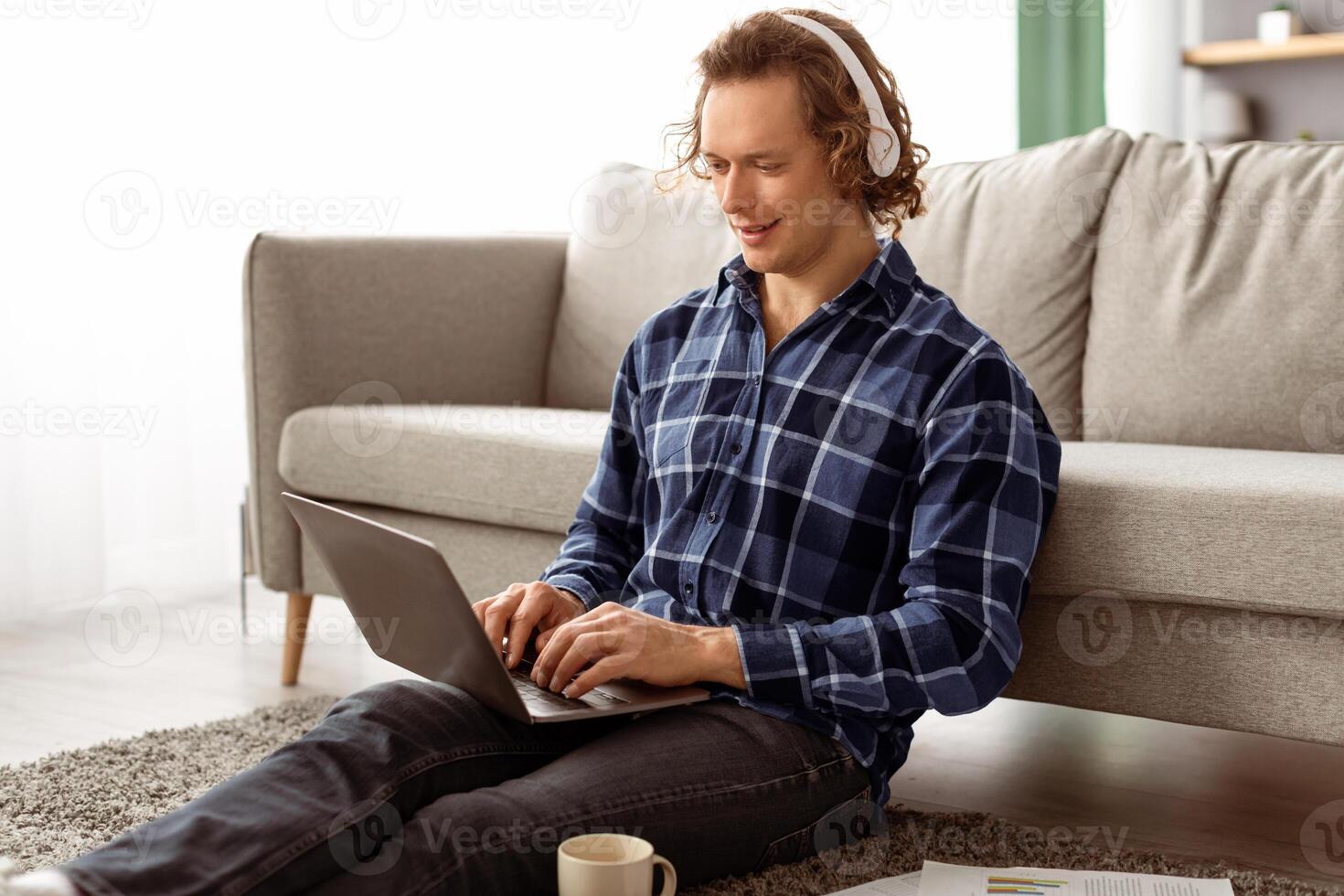 Smiling Man In Headphones Using Laptop Making Call Or Working Online Sitting On Floor At Home. Remote Job And Modern Communication, Internet And Technology Concept photo