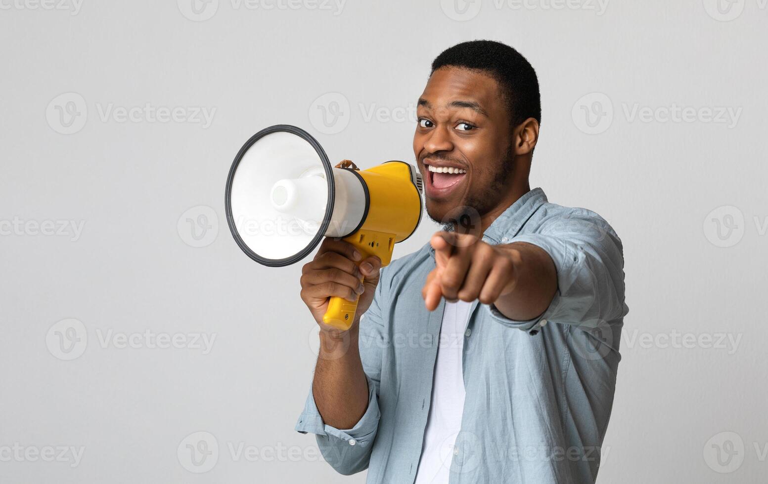 Positive african american guy shouting in megaphone and pointing at camera over grey background, copy space. Happy black man screaming with loudspeaker, cheering up, making advertisement photo