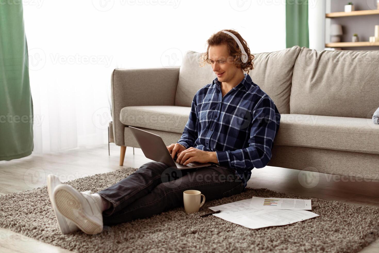 Happy Man Working On Laptop Wearing Headphones Browsing Internet Sitting On Floor At Home. Freelancer Guy Using Computer In Living Room. Distant Job, Internet And Technology photo